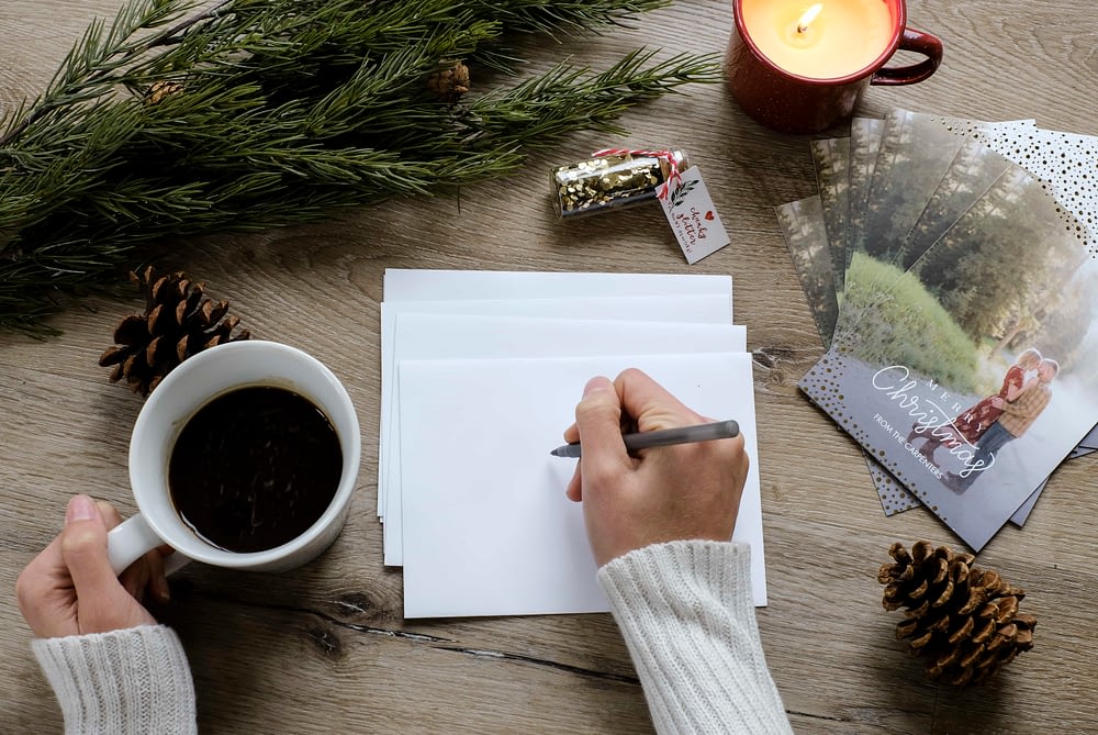 Overhead view of someone addressing envelopes next to a stack of personalized Christmas cards holding a coffee