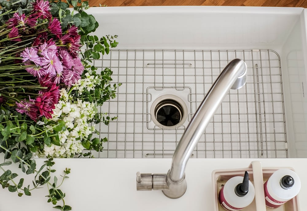 white sinkology farmhouse sink with metal grate in the bottom decorated with fresh flowers 