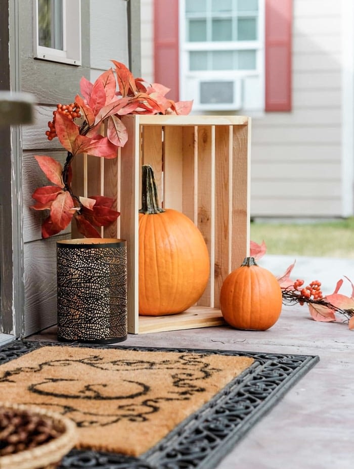 Fall front porch decor including a welcome mat, candle, pumpkins in a crate and a leaf garland