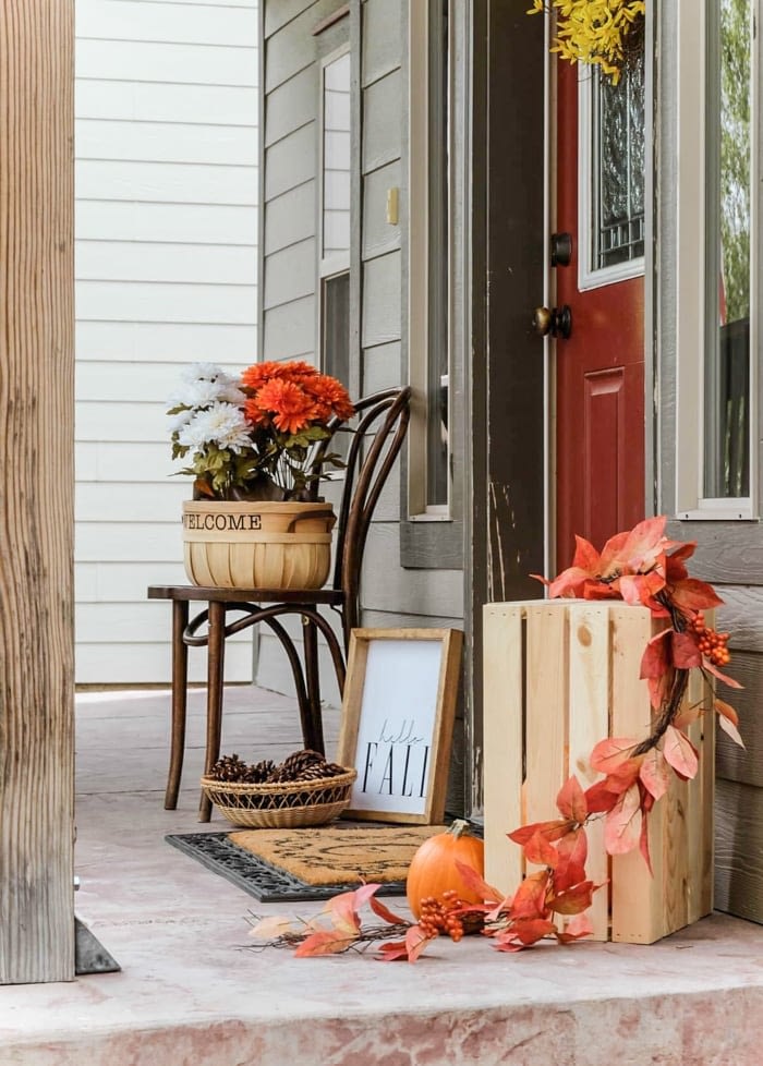 Fall front porch decor with orange and white flowers, pinecones in a basket and pumpkins in a crate