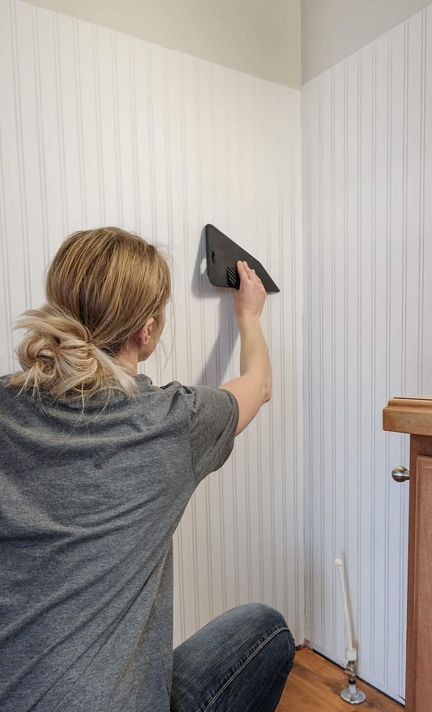 A woman using a wallpaper smoothing tool to smooth beadboard wallpaper onto a wall