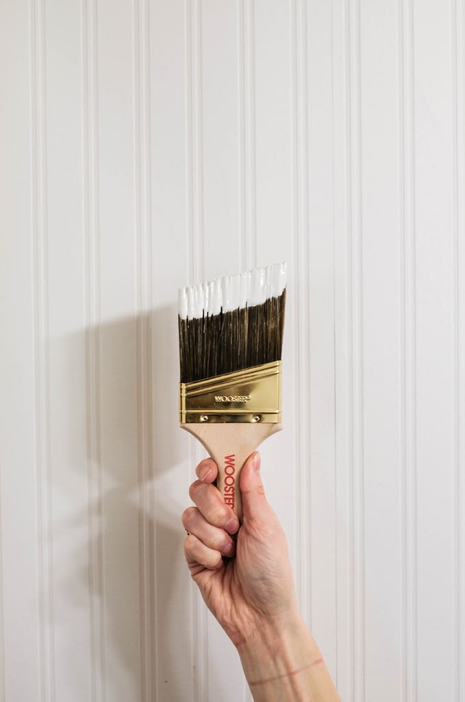 An angled paint brush dipped in white paint being held in front of white beadboad wallpaper