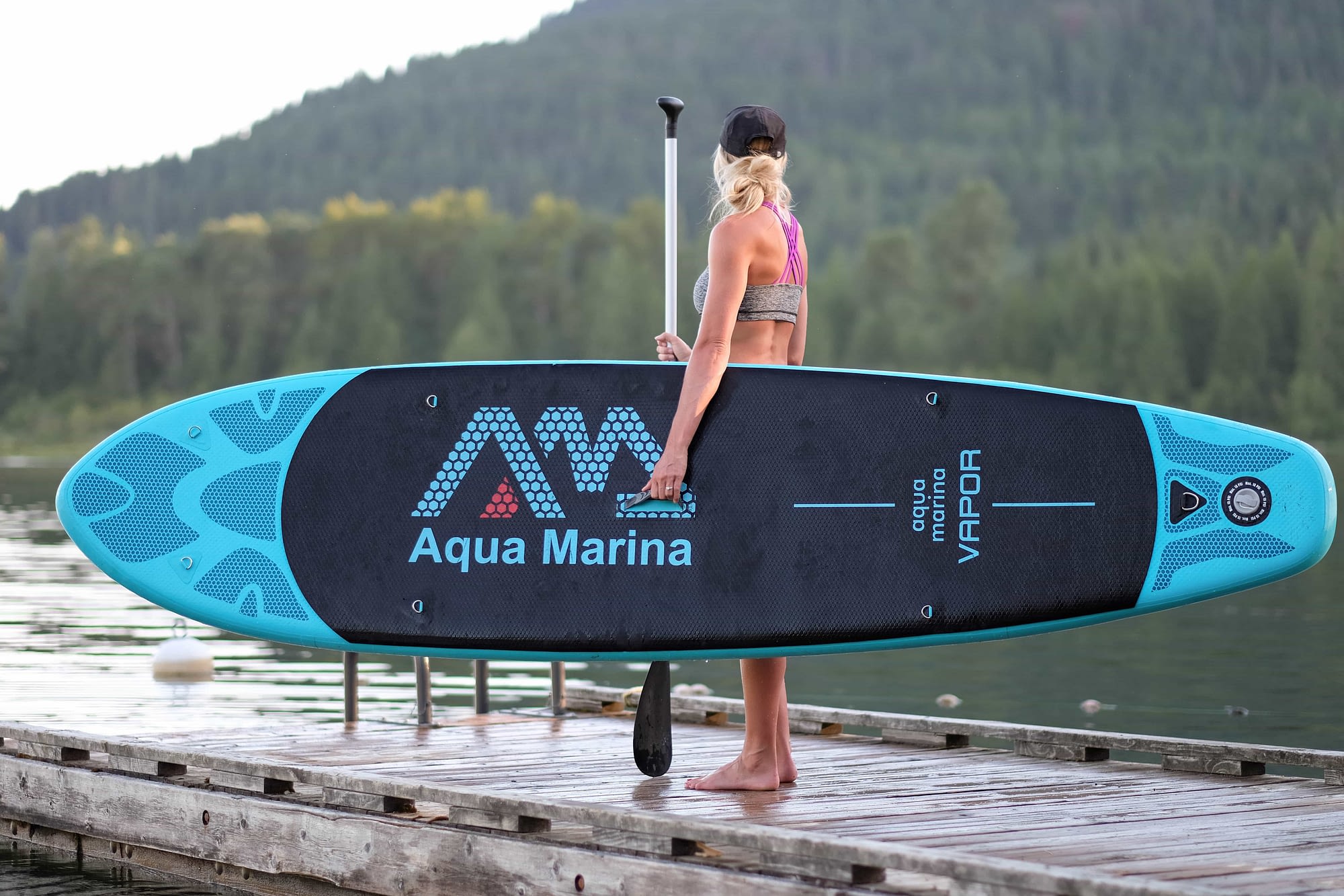 girl holding inflatable SUP standing on a dock looking over the lake